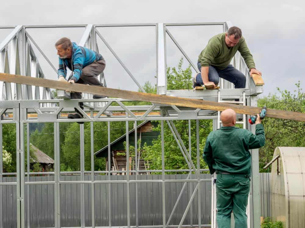 Trzech mężczyzn buduje metalową konstrukcję na świeżym powietrzu. Dwóch stoi na górze i mocuje drewnianą belkę, a trzeci podtrzymuje ją od dołu. W tle widać drzewa i ogrodzenie.